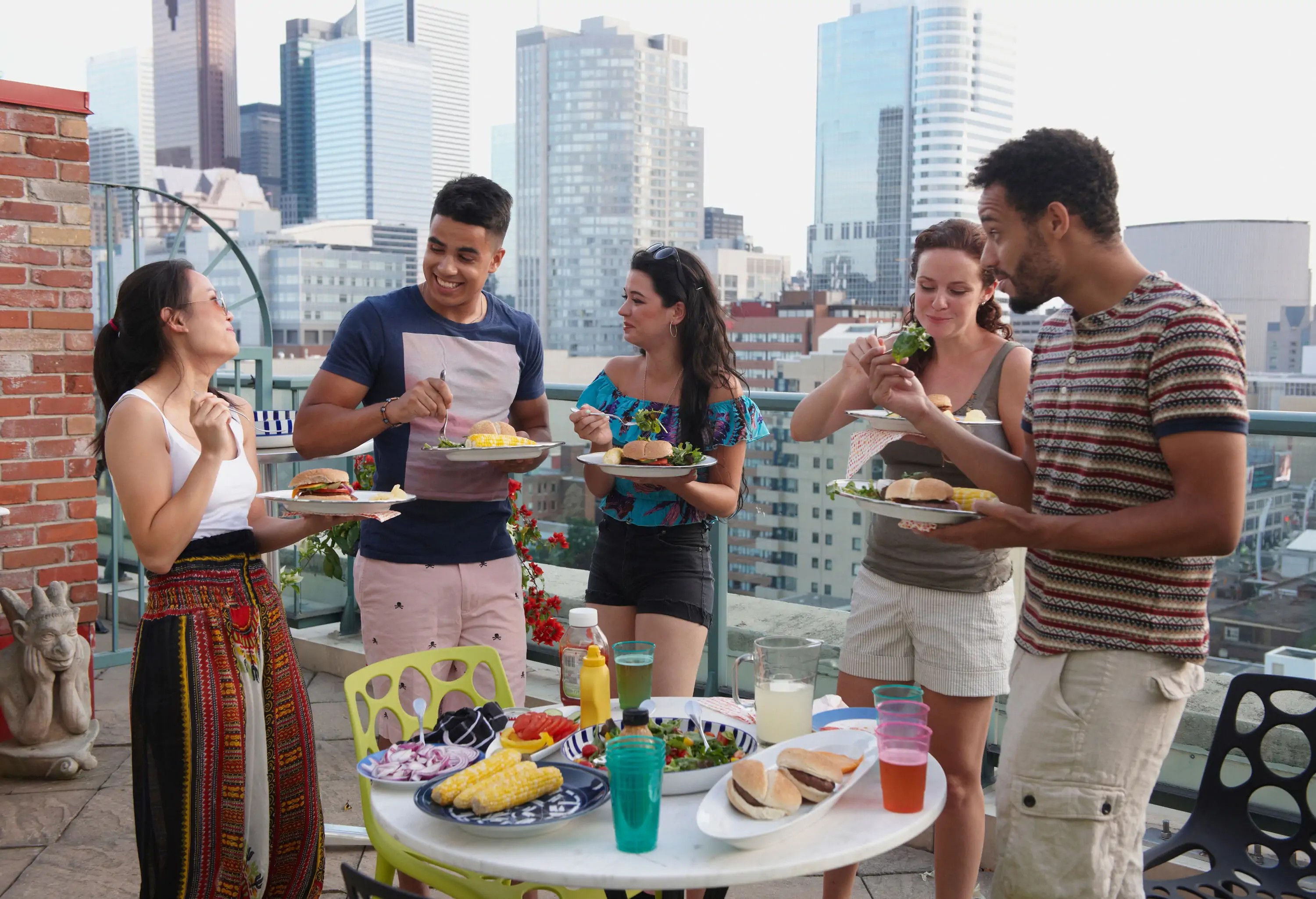 Group of friends having food and chatting whilst standing up on a rooftop, surrounded by tall skyscrapers