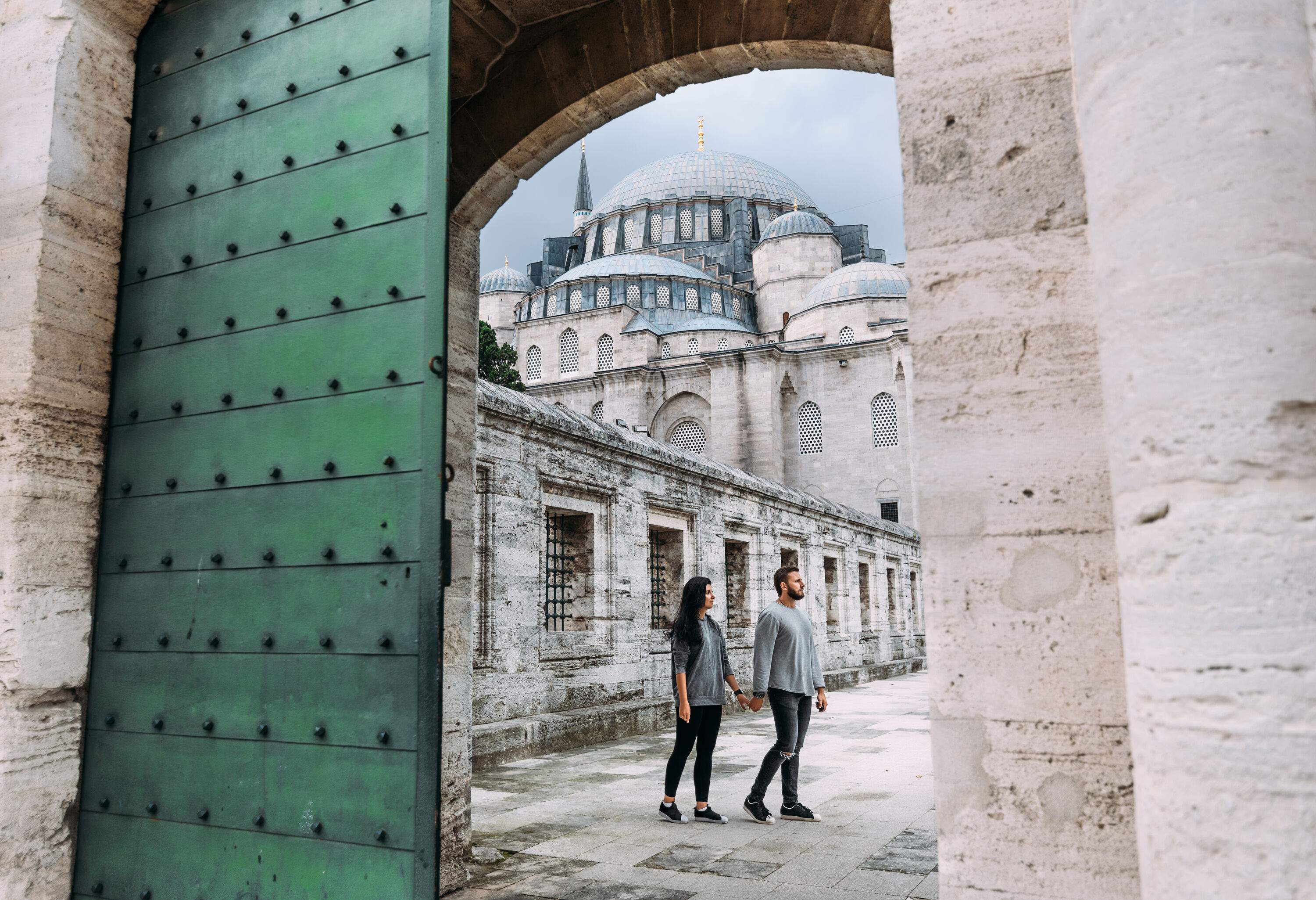 A couple holds hands while wandering in a mosque's courtyard guarded by a green arch entrance.