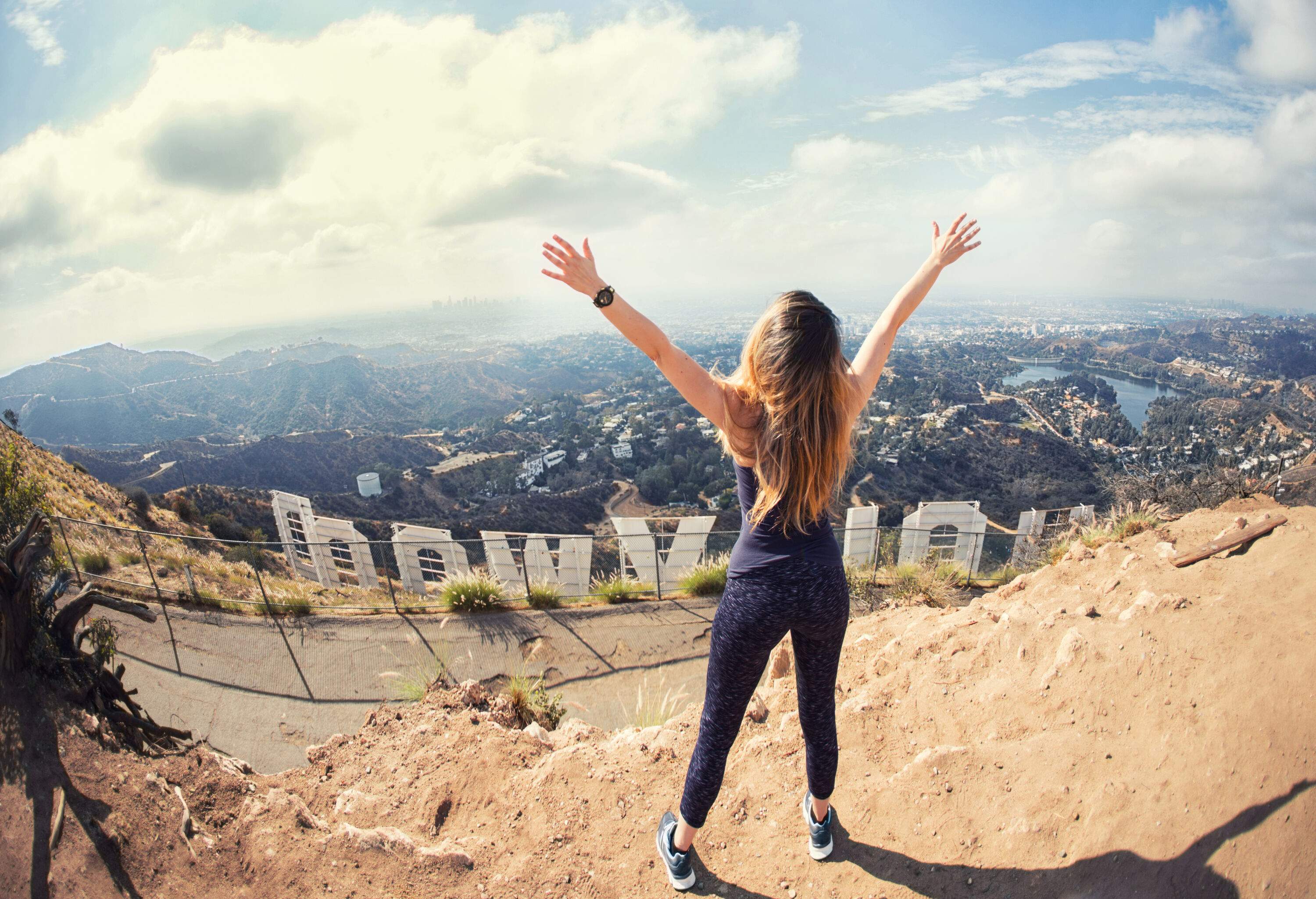 A woman spreads her arms as she stands on top of a Hollywood signage facing the residential hills.