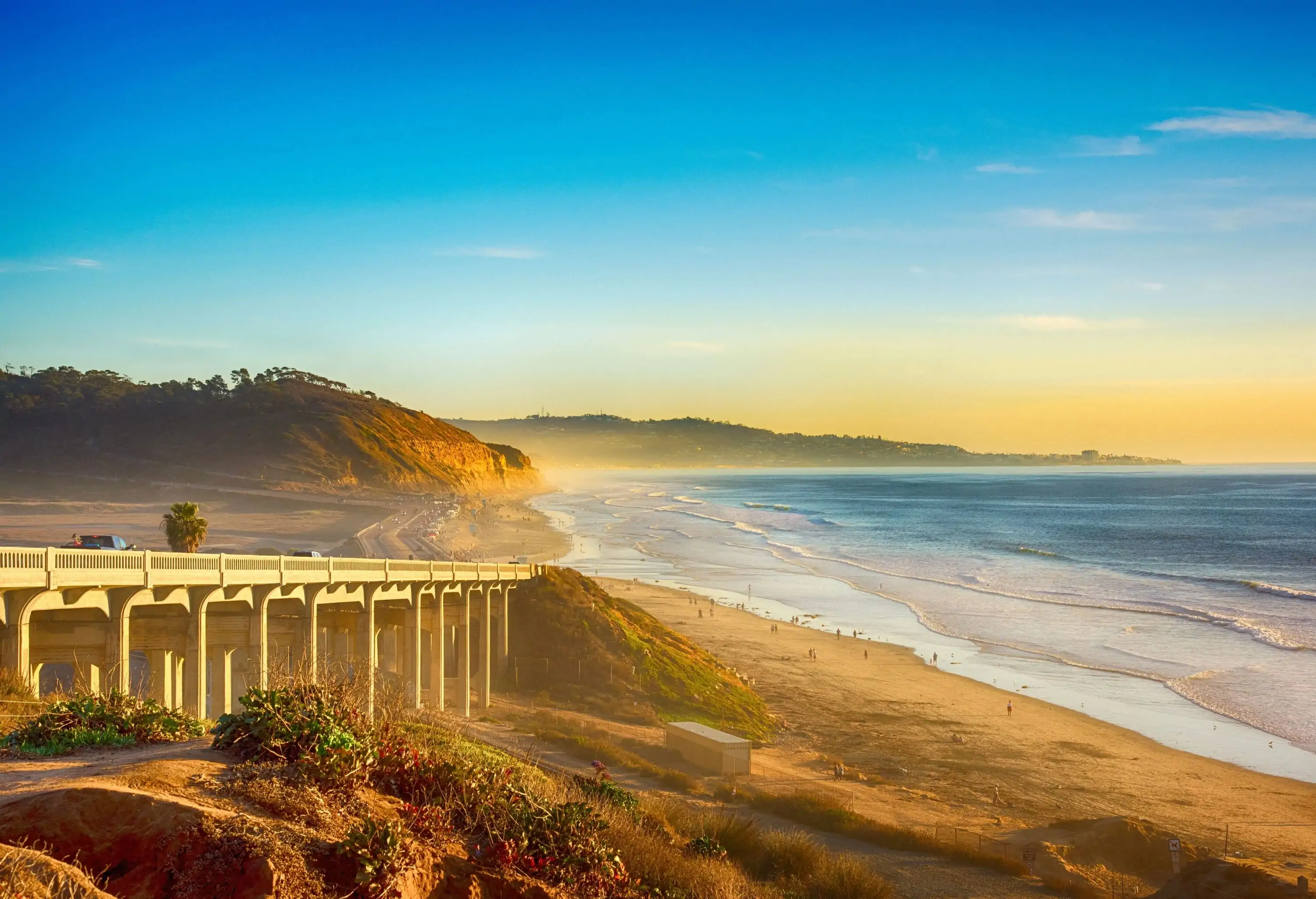 An elevated road bridge overlooks a beach with gentle waves during sunset.