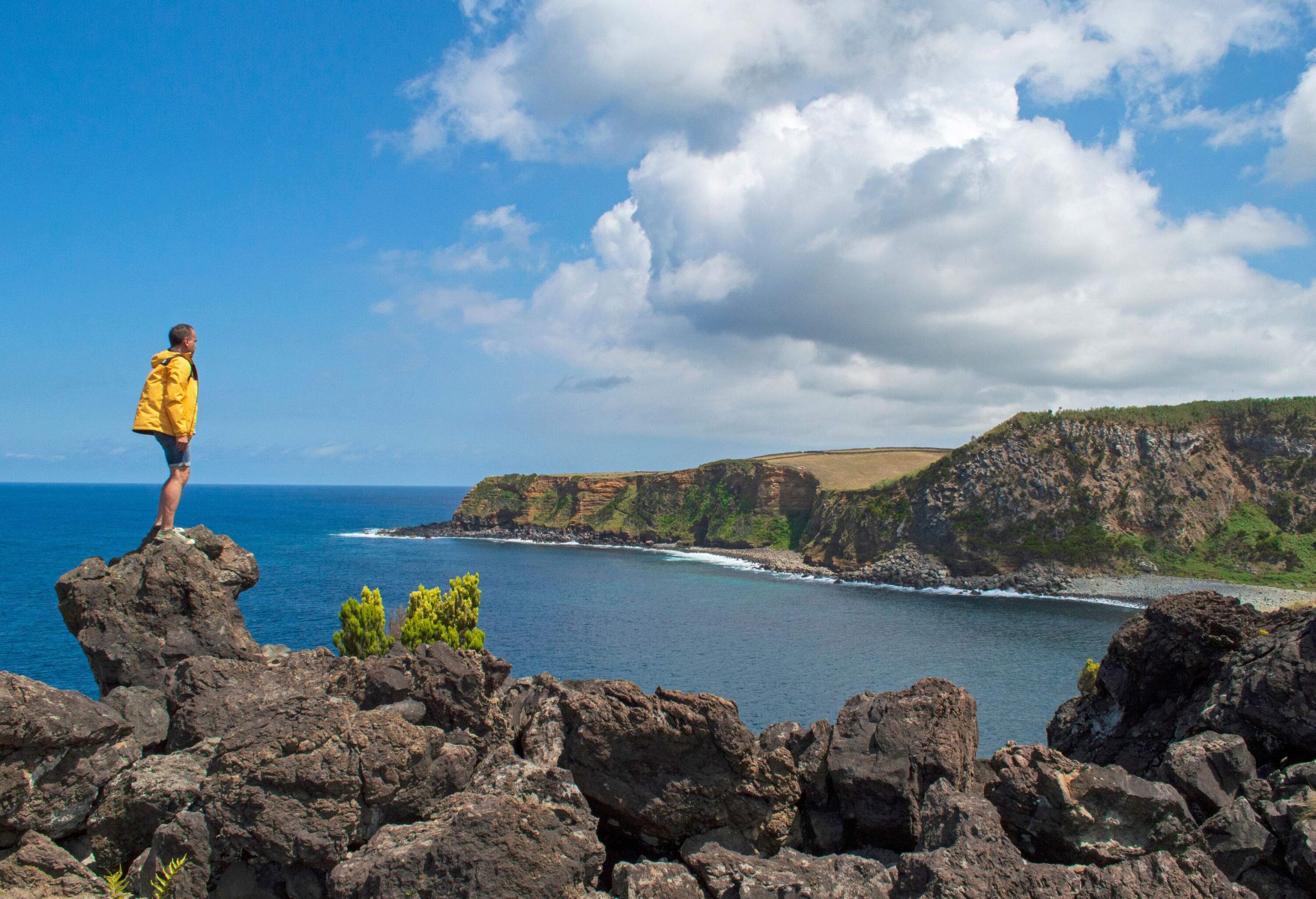 A man in a vibrant yellow jacket stands on a rock outcrop overlooking the vast ocean bounded by steep mountains.