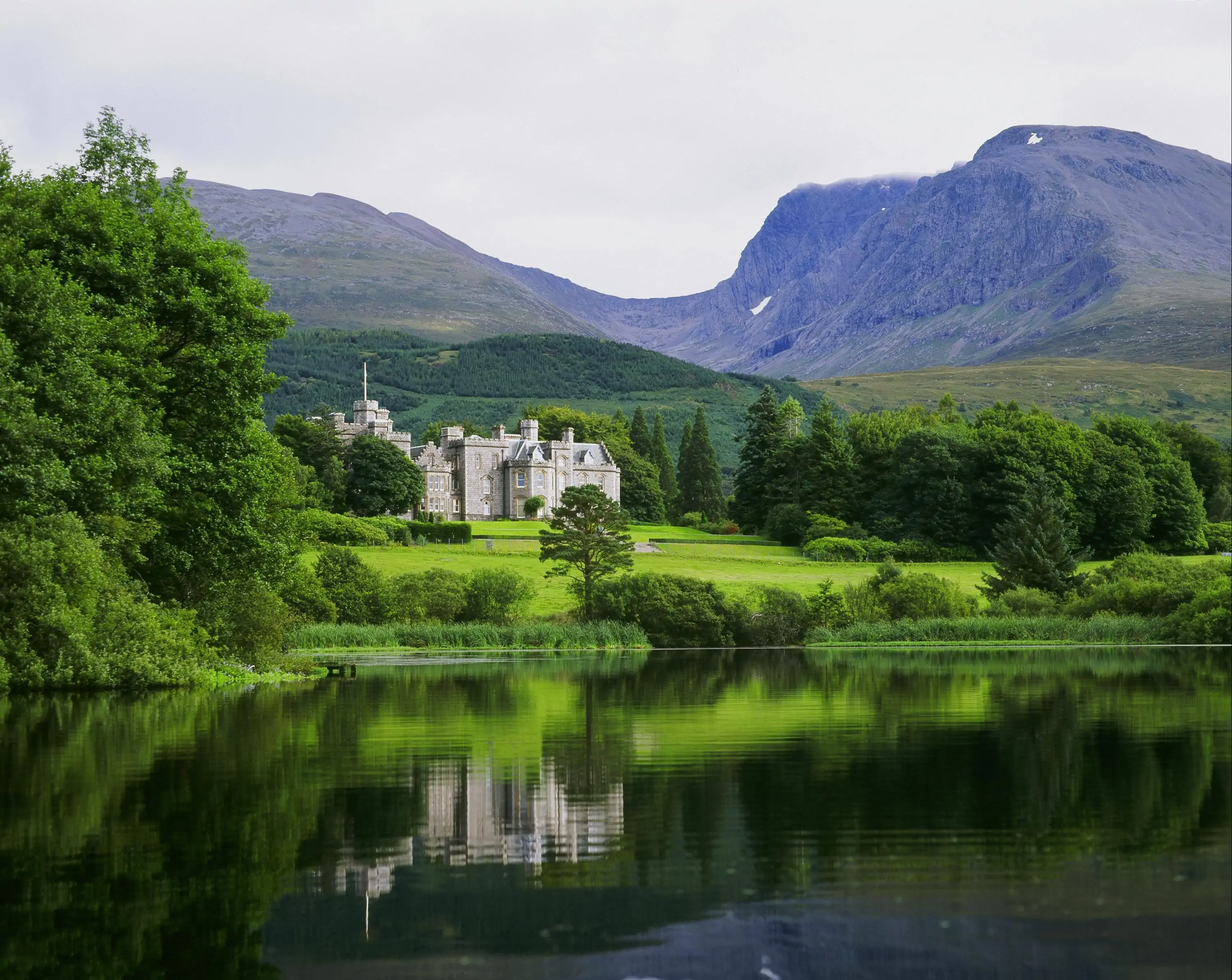 View of a castle reflected on a lake surrounded by green fields and mountains