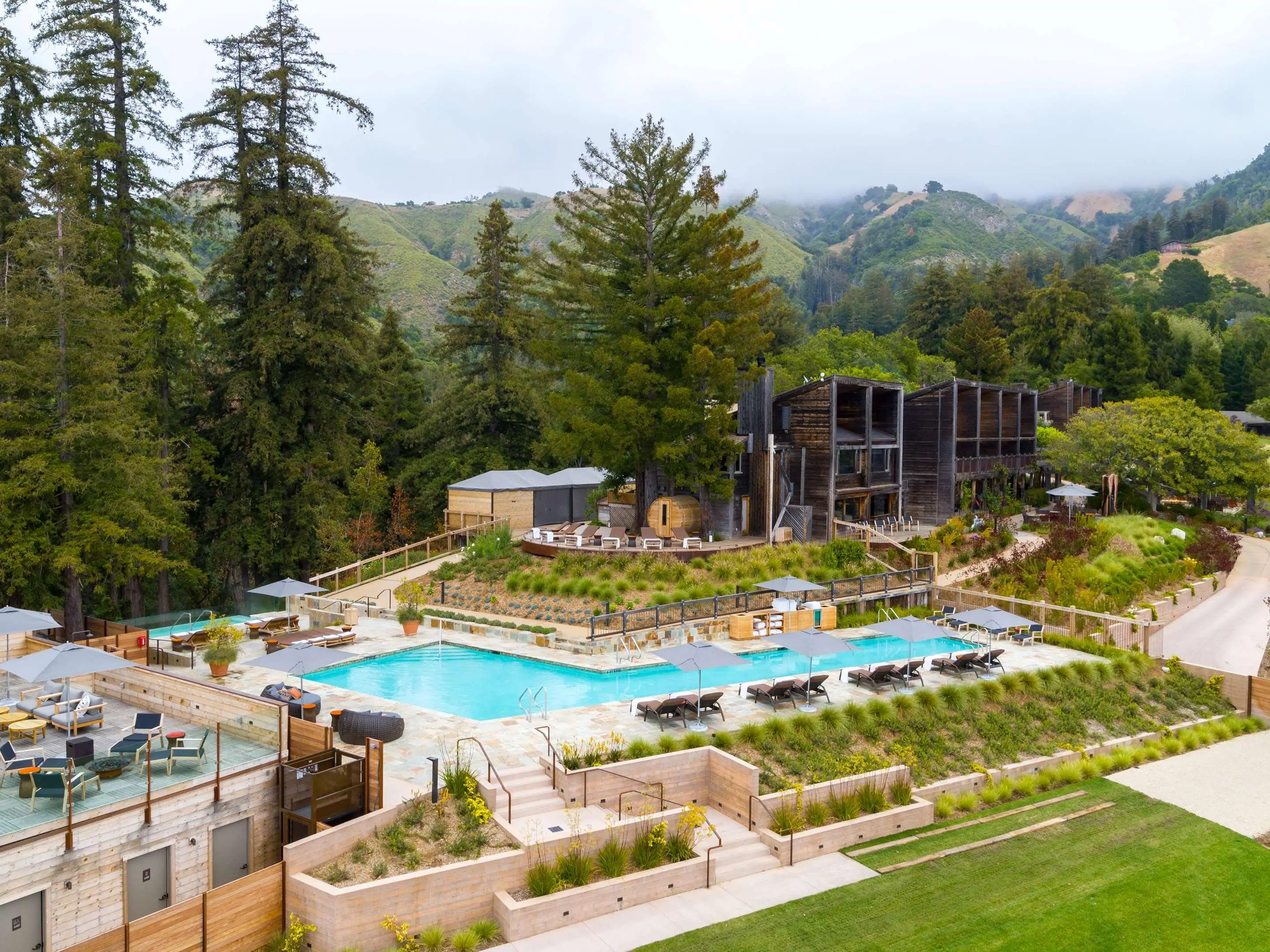 Resort pool area with loungers, surrounded by trees and hills under a cloudy sky.